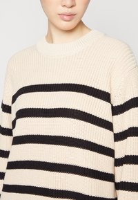 Beżowy sweter z dzianiny z czarnymi poziomymi paskami, prążkowaną fakturą, okrągłym dekoltem i delikatnym połyskiem. Zbliżenie na górną część klatki piersiowej.