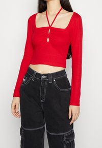 Kvinde iført en rød langærmet crop top med halterneck og nøglehulsudskæringer, parret med højtaljede sorte cargo-bukser med hvid syning.