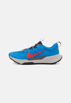 Baskets de sport bleues avec une tige en textile, logo central rouge, lacets marron et semelle contrastante grise et noire. Comprend une languette au talon.