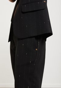 Černá pruhovaná saka s zlatými akcenty, obsahující jedno hrudní kapsu a štíhlý střih. Tkanina vypadá hladce s mírným leskem.