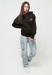Black hooded sweatshirt with front pocket, featuring a small floral logo. Paired with light blue cargo pants, textured denim, and white sneakers.
