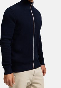 Marineblauer Zip-Pullover mit strukturiertem Strickmuster, Stehkragen und kontrastierendem Reißverschluss. Gerippte Bündchen und Saum sorgen für eine eng anliegende Passform.