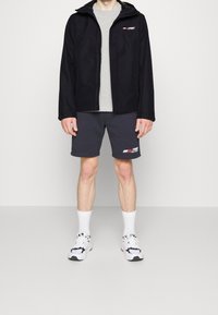 Sort vandtæt jakke med hætte, grå crewneck skjorte, mørkegrå shorts med logo, hvide sokker og hvide sneakers med sorte accenter.