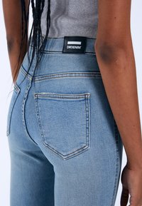 Light blue high-waisted jeans with a smooth denim texture, featuring a black label on the waistband and back pockets with contrasting stitching.