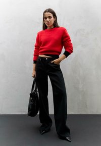 Maglione rosso tagliato con una finitura testurizzata e polsini a costine neri, abbinato a pantaloni neri a vita alta e gamba larga e una borsa nera.