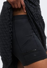 Gros plan d'une personne soulevant un vêtement noir à motifs pour révéler un short noir ajusté avec le logo JLindberg sur la zone de la poche.