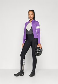 Lilla sykkeldress med hvit stripe, tettsittende svarte bib-shorts, svarte sko og en svart hjelm, som fremhever et strømmende, atletisk design.