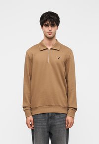 Καφέ μακρυμάνικη μπλούζα πόλο με φερμουάρ στον γιακά, μανσέτες με ριμπ ύφανση και μικρό λογότυπο στην αριστερή πλευρά, φορεμένη με σκούρο τζιν.