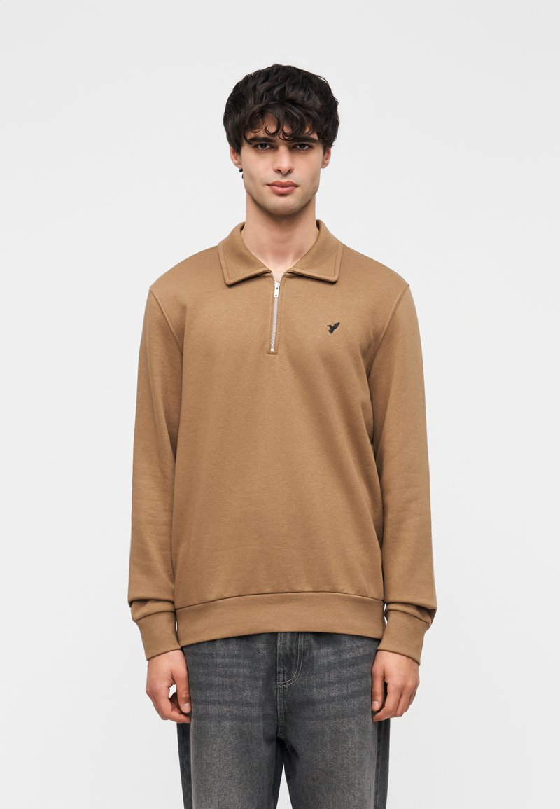 Καφέ μακρυμάνικη μπλούζα πόλο με φερμουάρ στον γιακά, μανσέτες με ριμπ ύφανση και μικρό λογότυπο στην αριστερή πλευρά, φορεμένη με σκούρο τζιν.