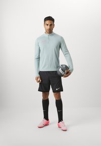 Lys blå langærmet sportstrøje med lynlås, sorte shorts, sorte sokker og lyserøde sportsko. Holder en grå Nike fodbold.