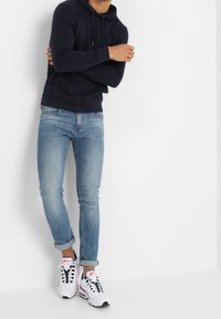 Sudadera con capucha azul marino con cordones, jeans entallados azul claro y zapatillas blancas con detalles en negro y rosa, de pie contra un fondo blanco.