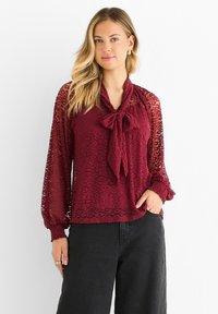 Burgundy blouse with sheer leopard print, a tied neck detail, and elasticized cuffs, paired with black high-waisted pants. Smooth texture.