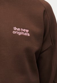 Brūns auduma džemperis ar rozā izšūtu tekstu "the new originals" krūtīs, redzamas plecu un augšdelma šuves.