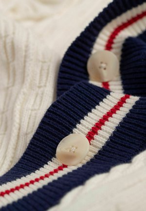 Strickjacke mit einer cremefarbenen Basis und einem marineblauen gerippten Kragen, akzentuiert mit roten und weißen Streifen sowie großen cremefarbenen Knöpfen.