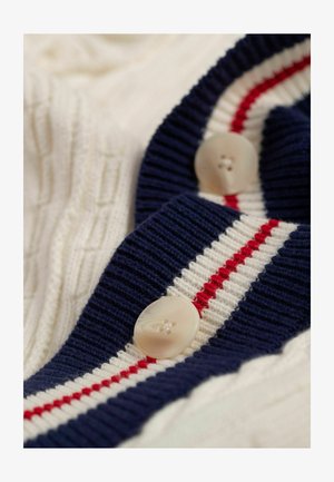 Strickjacke mit einer cremefarbenen Basis und einem marineblauen gerippten Kragen, akzentuiert mit roten und weißen Streifen sowie großen cremefarbenen Knöpfen.
