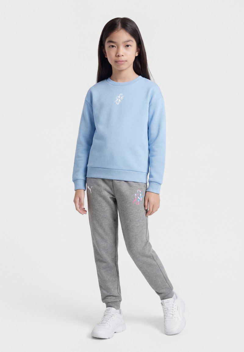 Mladá dívka stojící, oblečená v světle modré mikině, šedé tepláky s logy a bílých teniskách, na jednobarevném světlém pozadí.