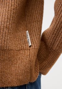 Pulover tricotat, cu nervuri, de culoare maronie, cu o textură moale și un eticheta Calvin Klein pe lateral. Designul prezintă o croială lejeră și manșete groase.