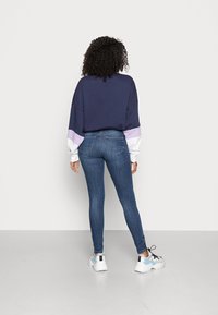 Sweatshirt oversized azul-marinho com mangas roxas e brancas, combinado com jeans justos azul e ténis brancos com detalhes azuis.