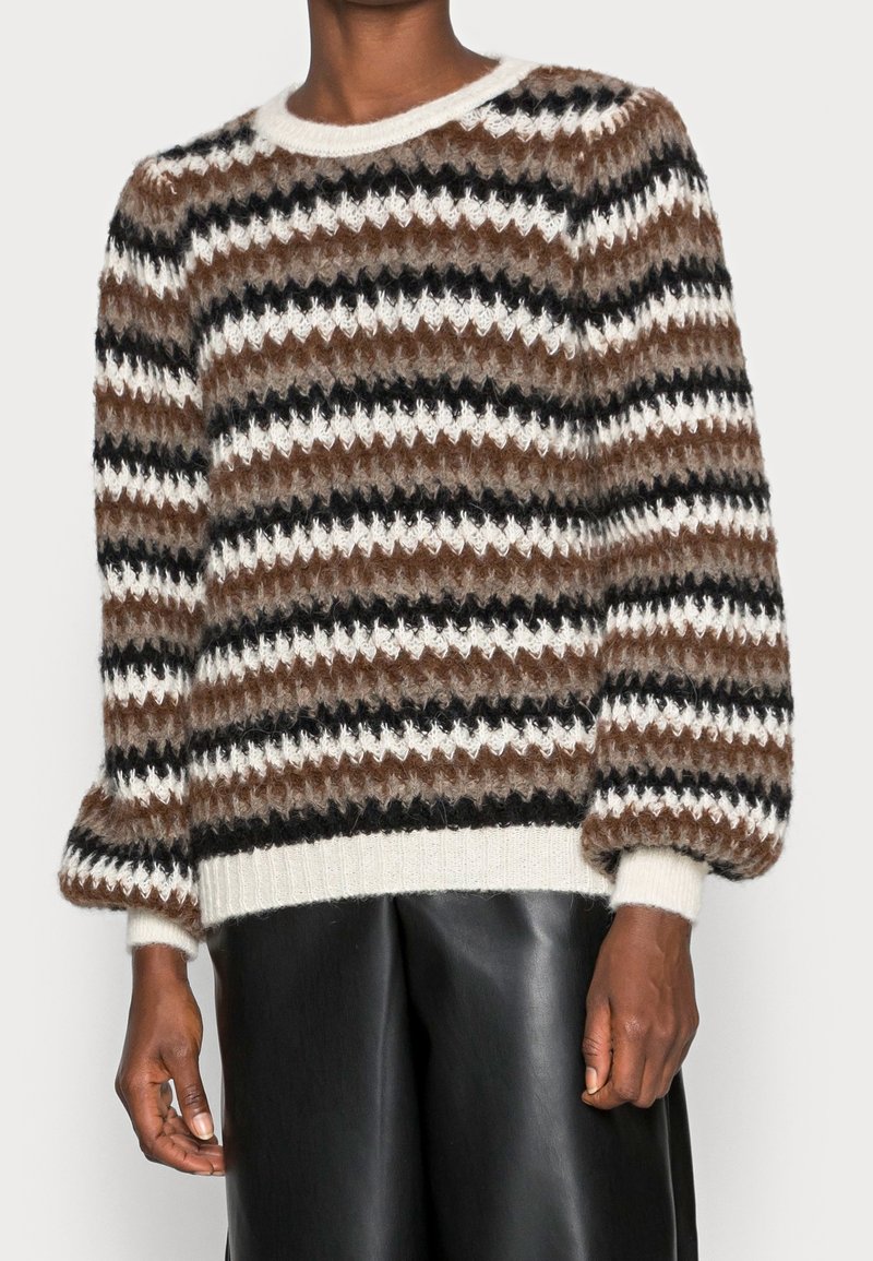 En modell som bär en randig, brun, svart och vit stickad tröja med puffärmar, tillsammans med svarta läderbyxor, vilket visar en modern stil.