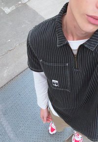 Mustreeritud must-valge lühikeste varrukatega shirt, millel on lukuga krae ja eesmise taskuga, kantud valge pikkade varrukatega särgi ja beežide pükste peal, seljas punased tennisetossud.