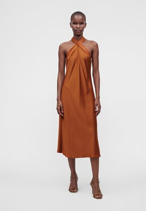 Femme portant une robe midi sans manches couleur rouille à col halter, avec des talons à brides à la cheville, debout sur un fond blanc uni.