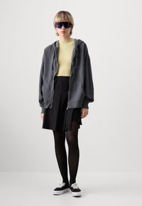 Šedá mikina na zip přes světle zelený top, černá skládací sukně, černé punčocháče a černé boty s bílou podrážkou. Velké fialové sluneční brýle.