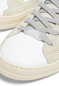 Gros plan sur une basket beige clair et blanche avec des lacets gris et une étiquette portant l'inscription "Project Sustainability" sur la zone des lacets.