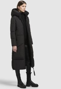 Manteau long noir en doudoune avec capuche, texture matelassée, fermeture éclair à l'avant et poches latérales, porté avec un pantalon en cuir noir et des bottes noires épaisses.