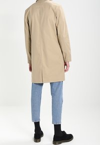 Cappotto beige con una silhouette dritta, maniche lunghe e uno spacco posteriore, abbinato a jeans azzurri a gamba affusolata e scarpe nere.