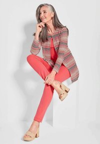 Bunt gemusterter Cardigan über einem korallenfarbenen Top und Hose. Beigefarbene Keilsandalen. Modell sitzt mit langen grauen Haaren und nachdenklichem Ausdruck.