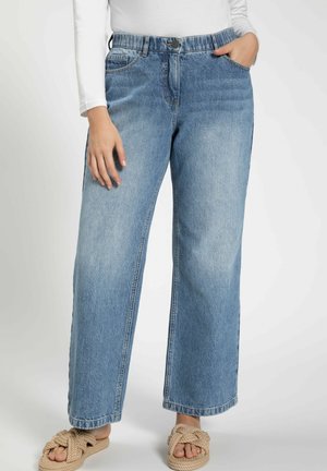 Personne portant un jean bleu clair à jambes larges avec un design cinq poches et des sandales tissées beige à enfiler, debout devant un fond uni.