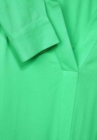 Tissu vert texturé en gros plan montrant le poignet de la manche longue et le détail de l'encolure en V d'un vêtement.