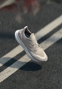 Chaussure de sport rose clair avec une tige en maille respirante, semelle blanche et détails texturés. Présente des lacets traditionnels et un logo.