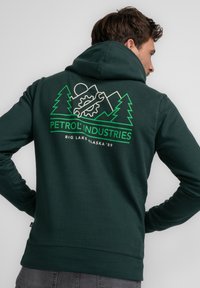 Sudadera verde oscura con un diseño gráfico de montañas, árboles y un engranaje. El texto dice "PETROL INDUSTRIES BIG LAKE ALASKA '89."