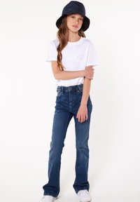 Kurze weiße T-Shirt mit einem Logo, dunkelblaue Jeans und ein marineblauer Bucket Hat, stehend mit verschränkten Armen vor einem hellen Hintergrund.