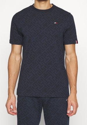 Mann trägt ein dunkel gemustertes Kurzarmhemd und passende Shorts mit kleinen roten, weißen und blauen Logodetails.