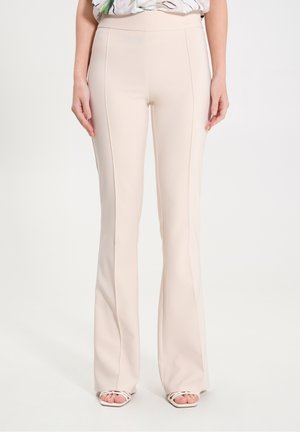 Kvinde iført beige højtaljede viddebukser og hvide stropper med åbne tå-sandaler, stående med hænder afslappet ned langs siderne.