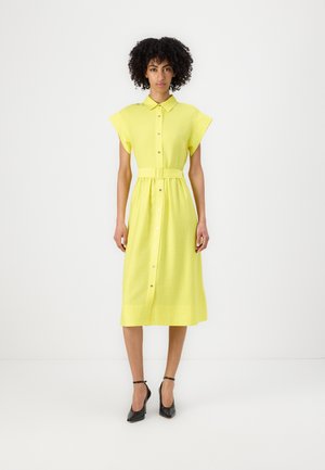 Vestito giallo con bottoni, maniche a cappotto corte, con cintura in vita. Realizzato in tessuto texture, arriva a metà polpaccio, abbinato a tacchi neri.
