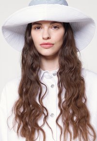Chapéu branco de aba larga com um design texturizado e uma faixa riscada em azul marinho, combinado com uma camisa branca com detalhes em botão e ondas suaves no cabelo.