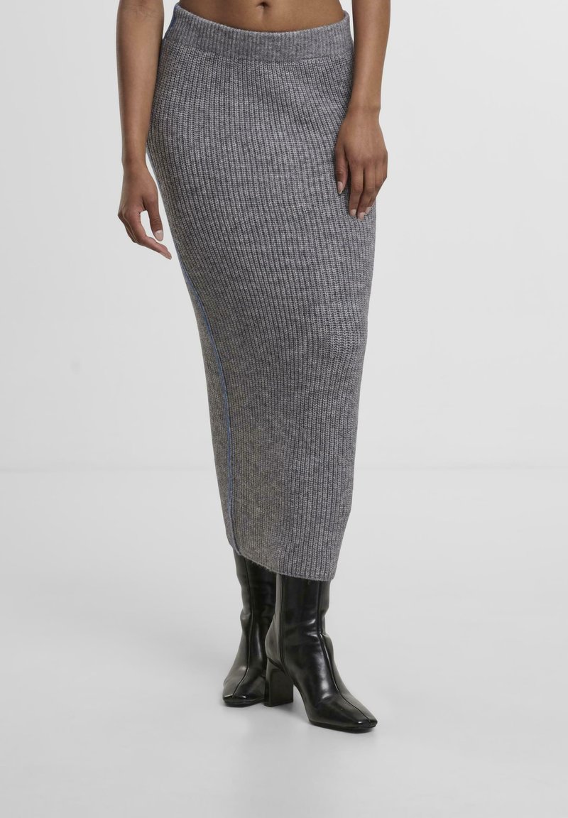 Gestrickter grauer Midirock mit geripptem Muster, versehen mit einer seitlichen Akzentuierung aus blauer Naht. Kombiniert mit schwarzen High-Heels.