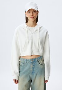 Haut en hoodie blanc coupé avec fermeture éclair à l'avant, cordon de serrage et deux poches ; associé à un jean bleu clair légèrement décoloré. Casquette blanche.
