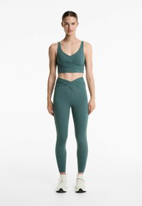 Atletisk set i mörkgrön, som består av en omslutsande sportbh och hög midja leggings med snurrade detaljer. Tillverkad av ett slätt tyg.