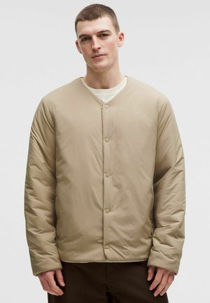 Mężczyzna ubrany w beżową, pikowaną kurtkę na guziki, na białej koszuli, stojący na jednolitym jasnym tle.