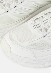 Hvite joggesko med mesh-paneler, polstrede såler og reflekterende detaljer. Logo trykt på siden, med teksturerte mønstre over hele skoen.