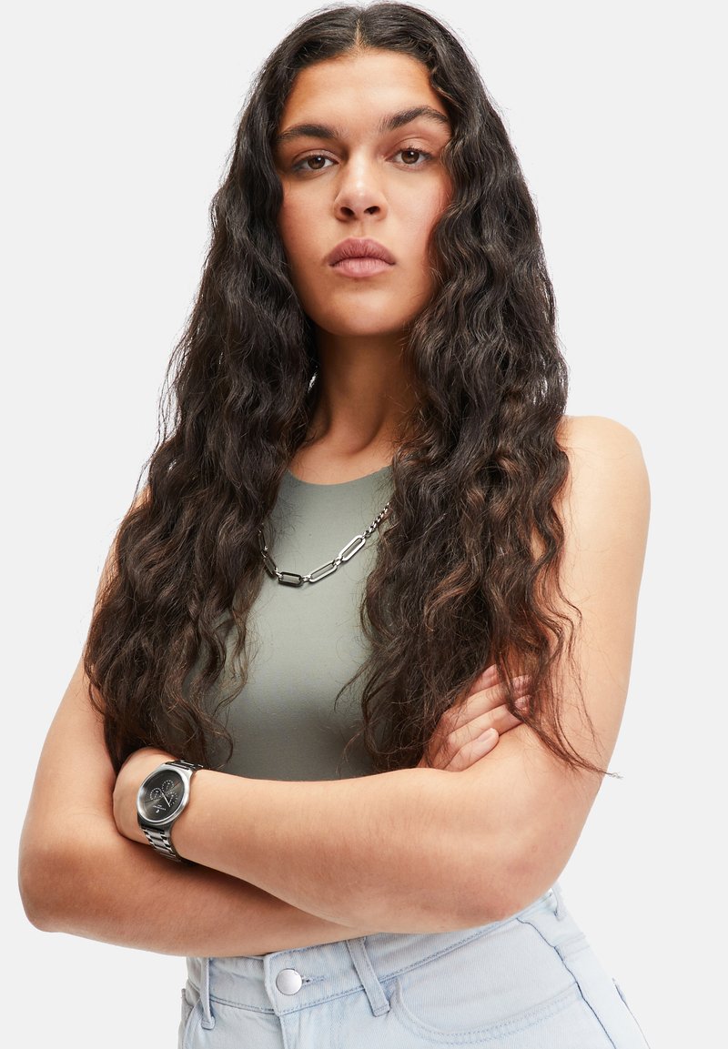 Silberne Armbanduhr, grünes ärmelloses Oberteil, große Kettenhalskette. Das Model hat langes, gewelltes Haar, die Arme verschränkt, trägt hellblaue High-Waist-Jeans.