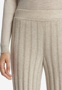 Beżowe rypsowe spodnie dzianinowe o miękkiej fakturze. Posiadają szeroki pas i pionowe paski dla dodatkowego detalu. Częściowo widoczna ręka.