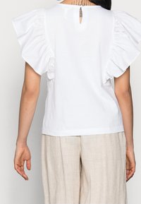 Top blanco de manga corta con mangas con volantes, abertura en la espalda con forma de llave y un corte relajado. Combinado con pantalones sueltos de color beige claro.
