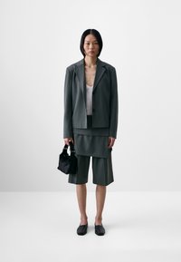 Dark green tailored blazer with two buttons, paired with a matching layered shorts set. Accessories include a small black fur handbag and black shoes.