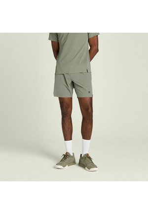 Mann trägt olivgrüne Sportshorts, passendes Shirt, weiße Kniesocken und olivgrüne Sneaker, steht vor einfachem Hintergrund.