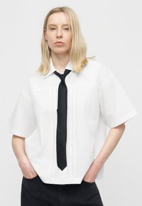 Νεαρή γυναίκα με ξανθά μαλλιά έως τους ώμους φοράει άνετο λευκό κοντομάνικο πουκάμισο και μαύρη γραβάτα, με τα χέρια στις τσέπες ενός ζευγαριού μαύρων παντελονιών.